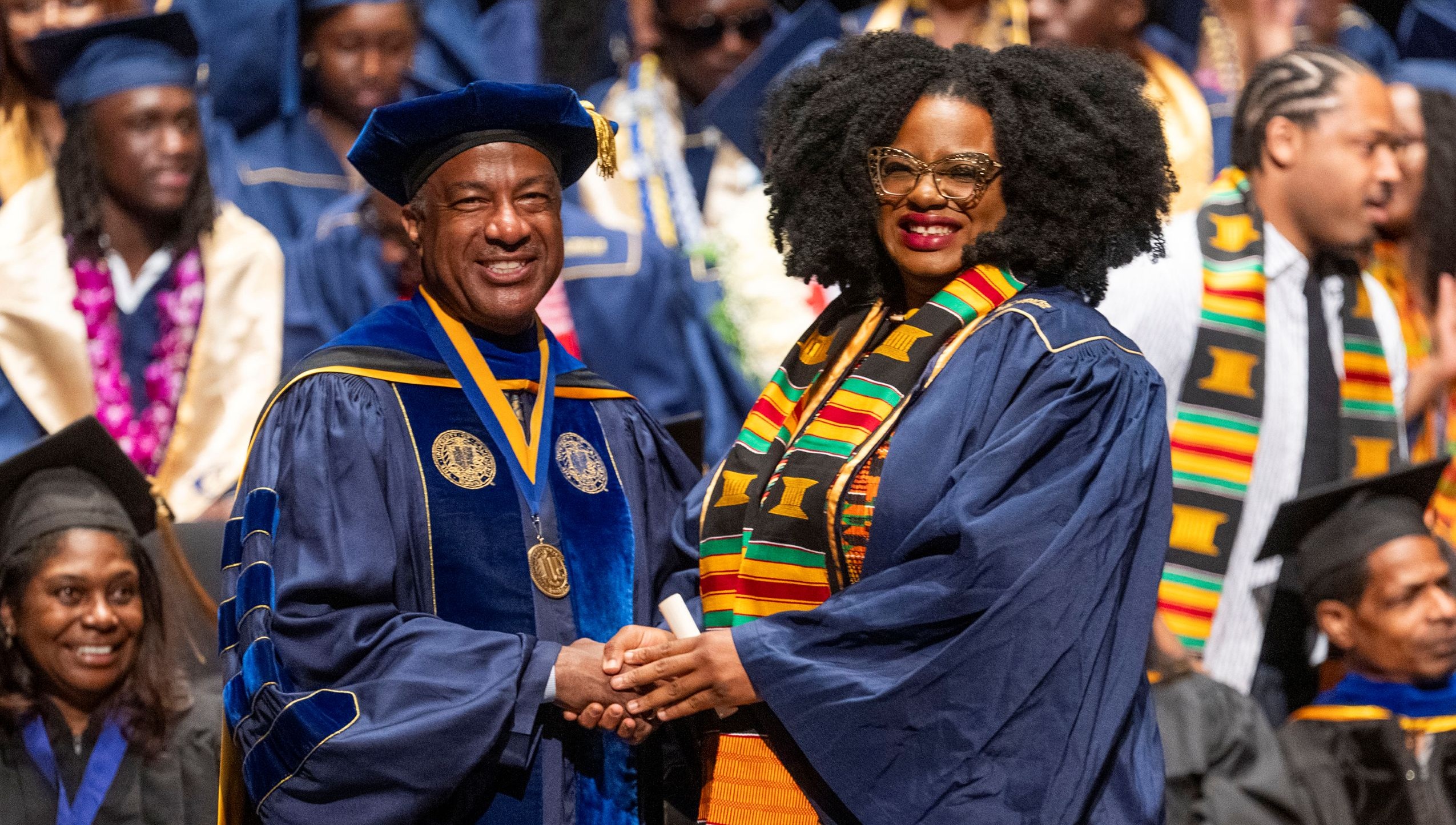 Tanika Baptiste on stage in regalia at Black graduation shaking hands with Chancellor May